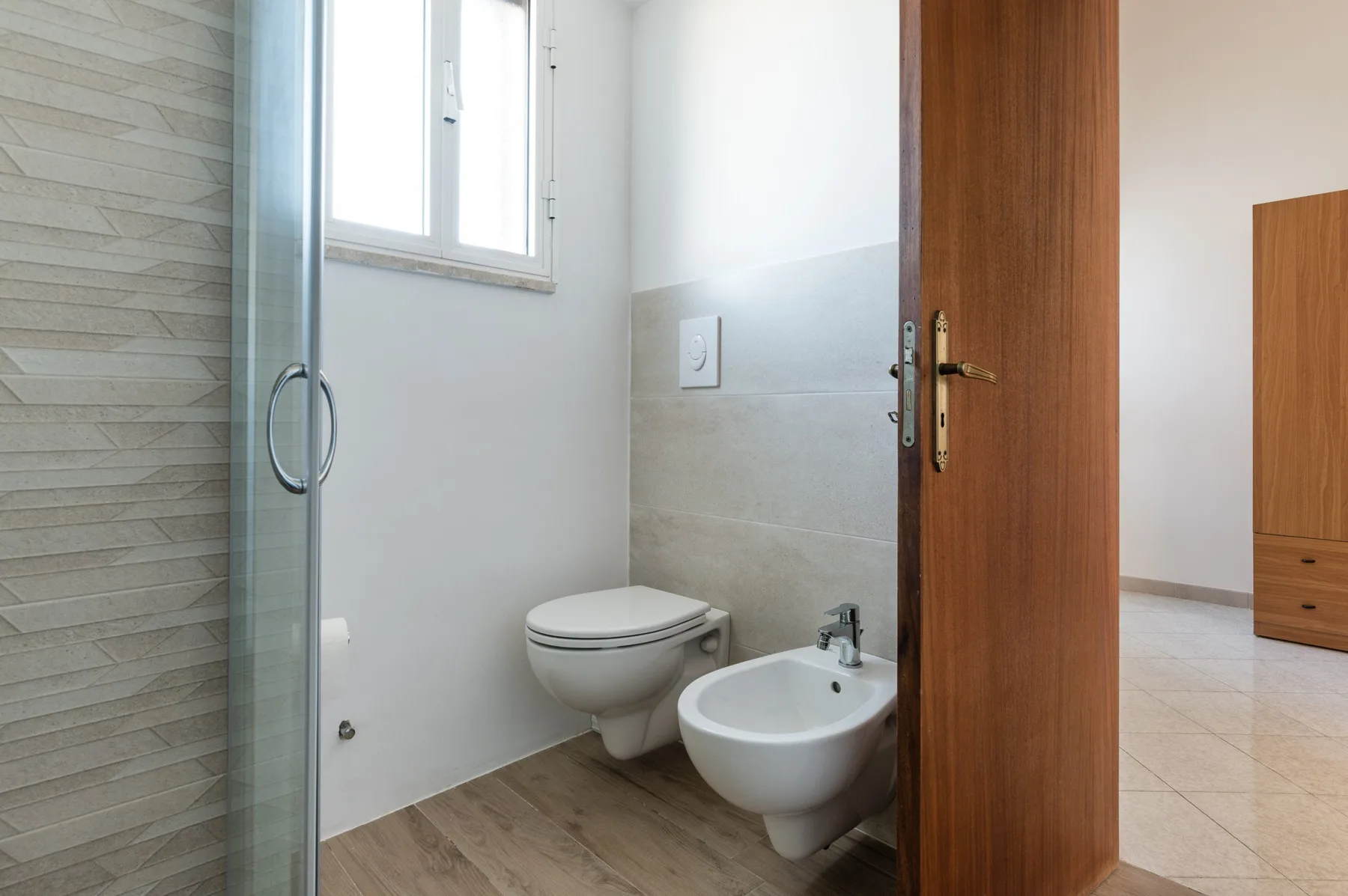 A bathroom with a glass shower, toilet, and bidet beside a wooden door.