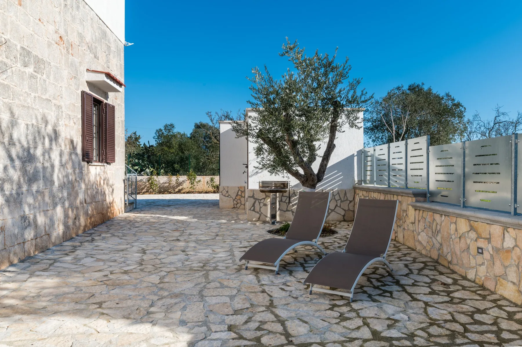 A stone-paved outdoor patio has two lounge chairs beneath a tree.