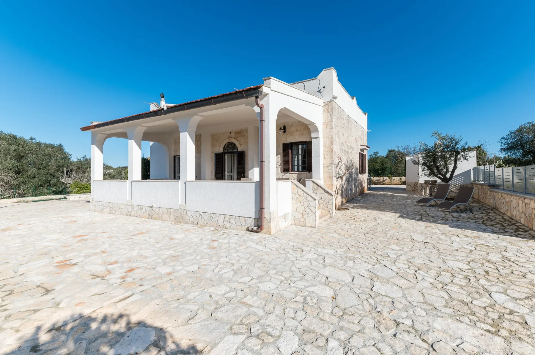 A white house with a covered porch stands beside a broad stone-paved courtyard.