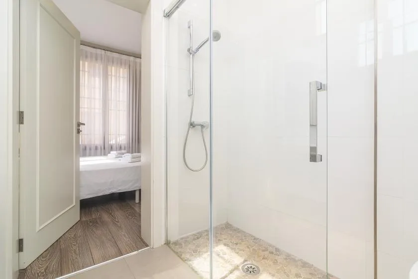 Un baño luminoso con una ducha de vidrio y un dormitorio visible a través de la puerta abierta.
