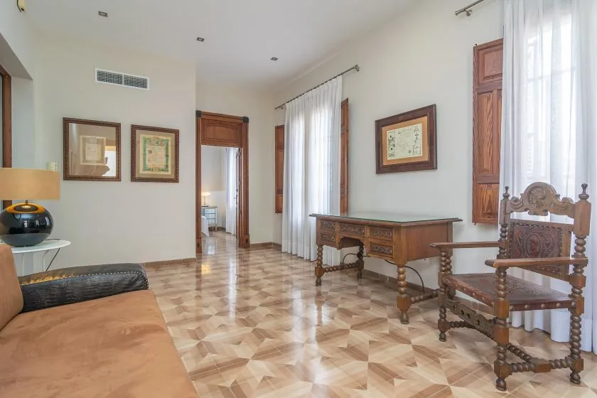 Una sala de estar luminosa con asientos de madera y un escritorio junto a ventanas altas.