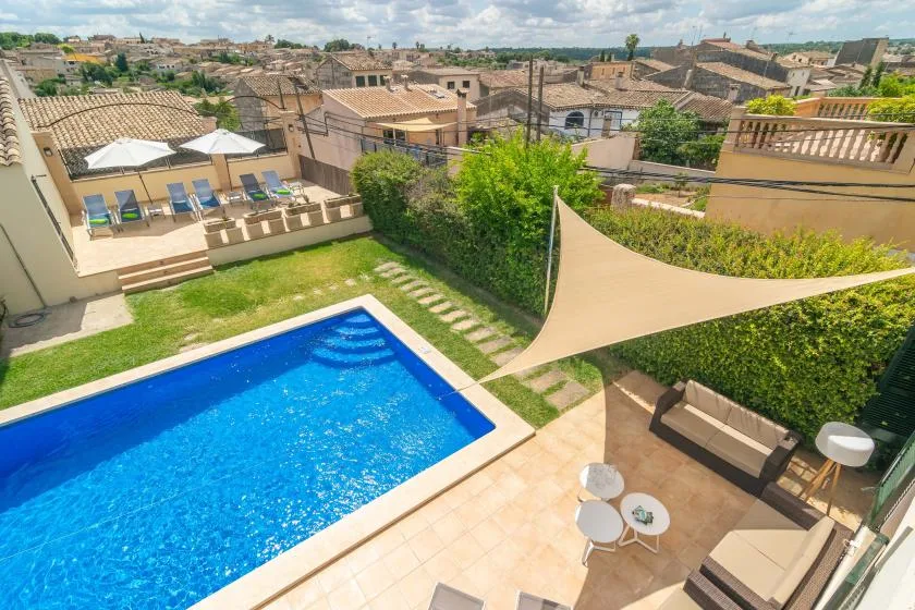 Una piscina exterior y un patio están junto a un césped bajo una vela de sombra.