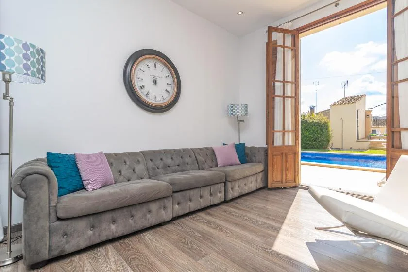 Una sala luminosa con un sofá gris largo junto a puertas abiertas hacia una piscina.