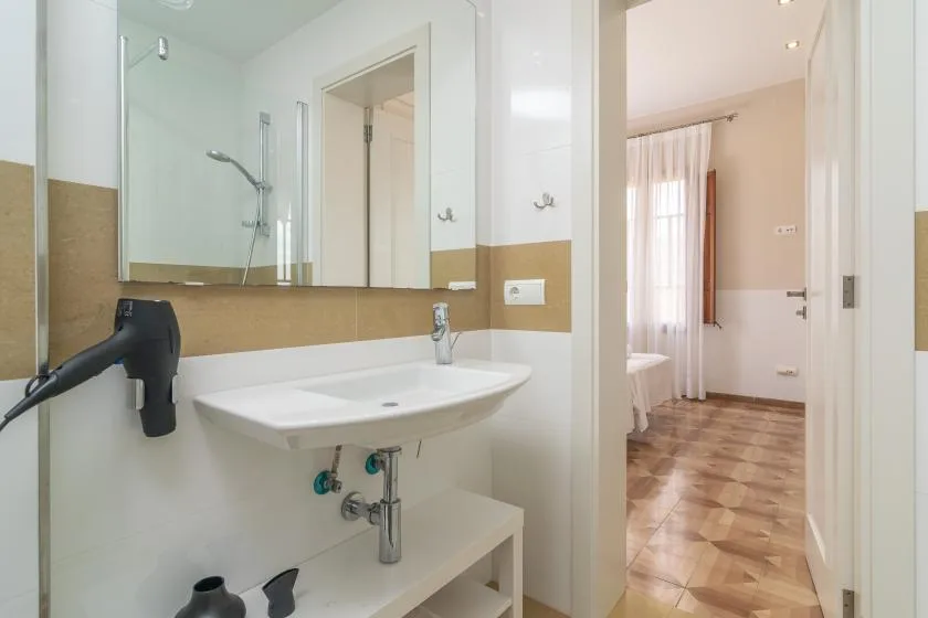 Un baño con lavabo y ducha, abierto a una habitación con una ventana con cortinas.