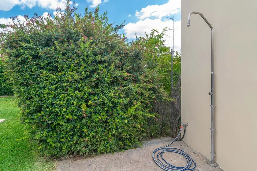 Una ducha exterior junto a un seto frondoso y una zona pavimentada.