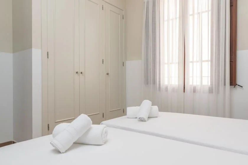 Un dormitorio luminoso con camas blancas y cortinas transparentes en la ventana.