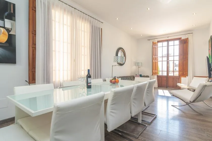 Un comedor luminoso con una mesa de vidrio y asientos blancos junto a ventanas altas.