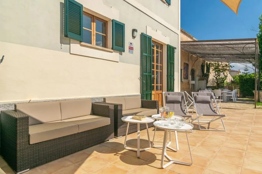 Un patio soleado con asientos y ventanas con contraventanas verdes junto al edificio.