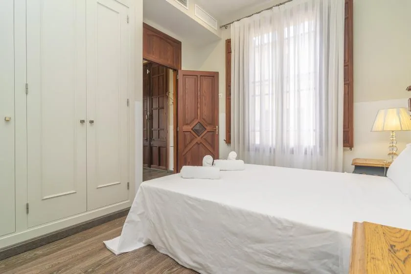 Un dormitorio con una cama junto a una ventana alta con cortinas translúcidas.