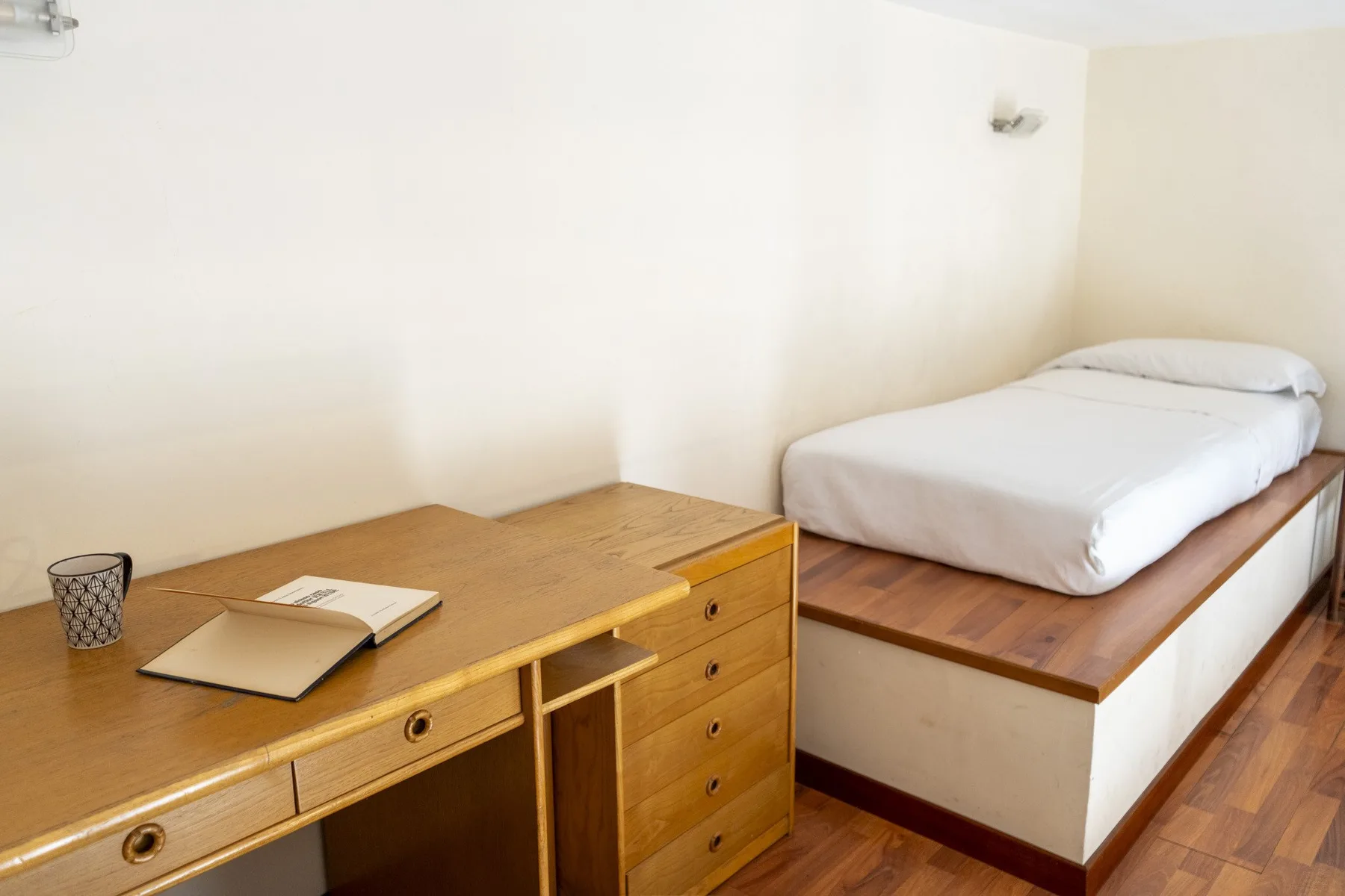 A bedroom with a wooden desk beside a bed with white bedding.