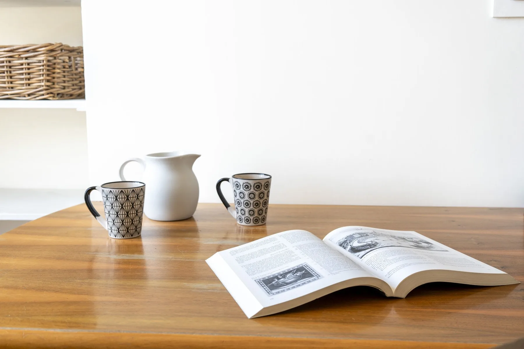 A wooden table holds an open book, two patterned cups, and a white pitcher.