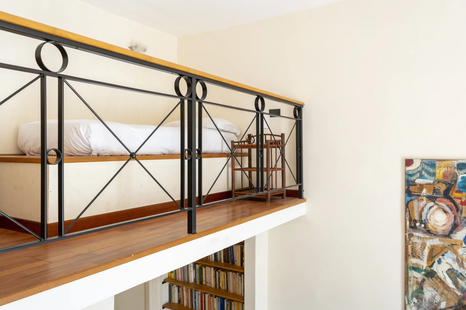 A raised sleeping area with a bed and black metal railing overlooks a bookshelf.