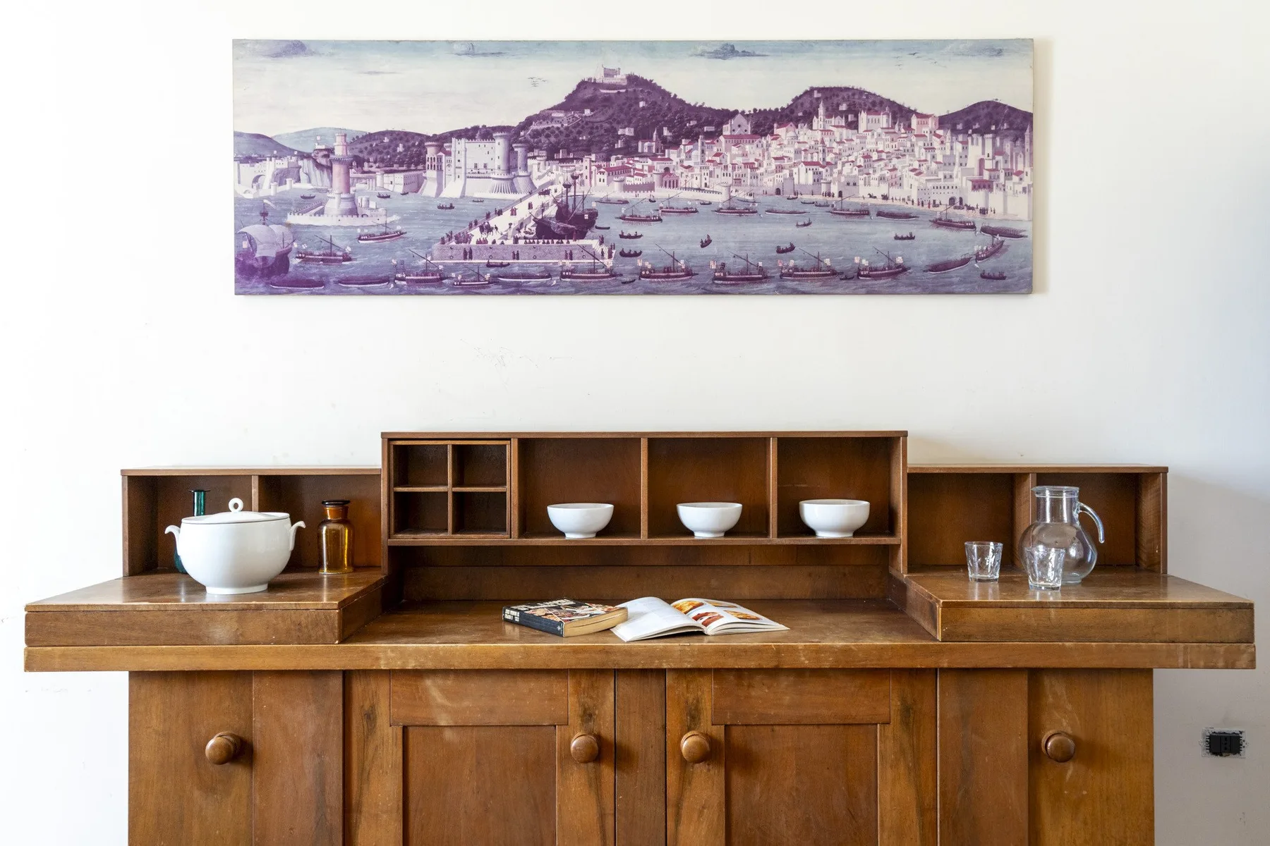 A wooden sideboard stands against a white wall beneath a framed harbor print.
