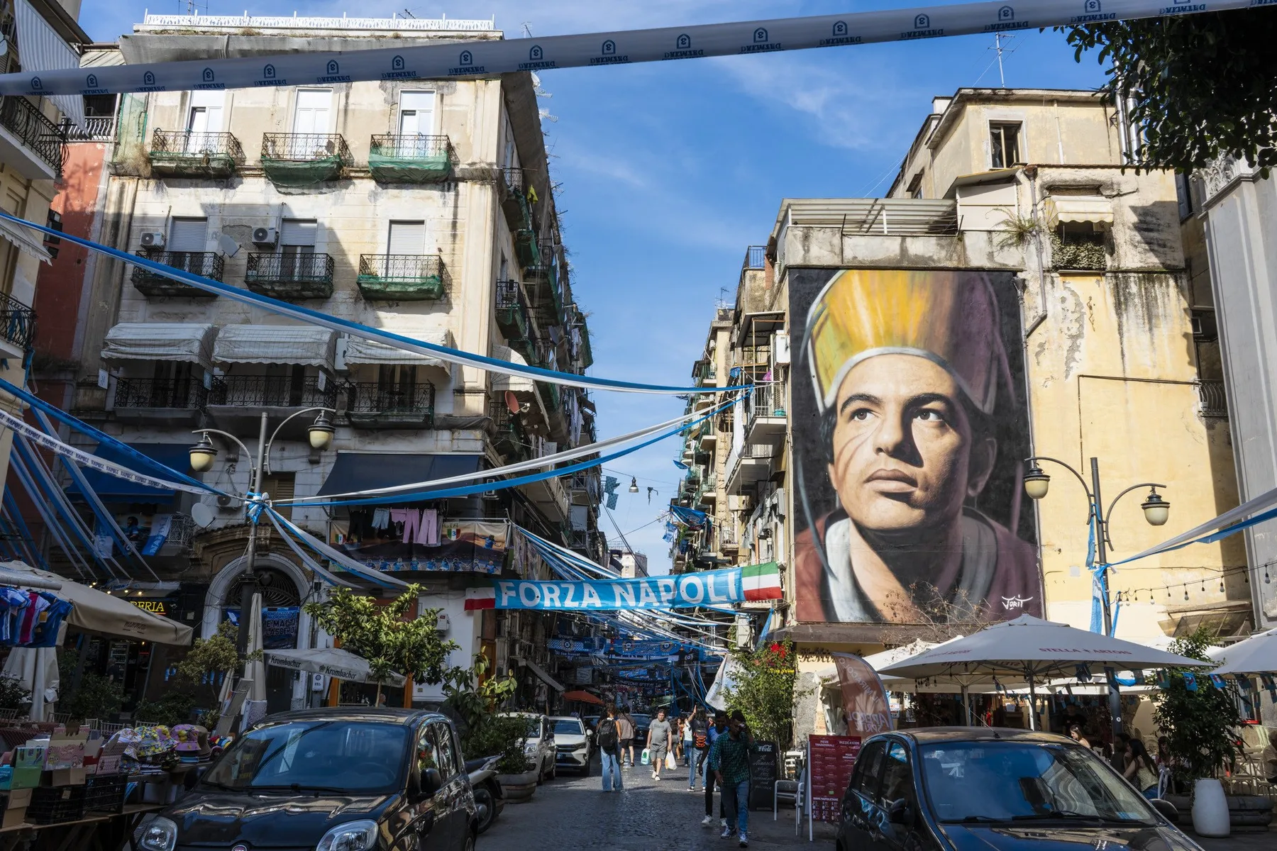 A narrow city street lined with buildings, parked cars, and a large mural.