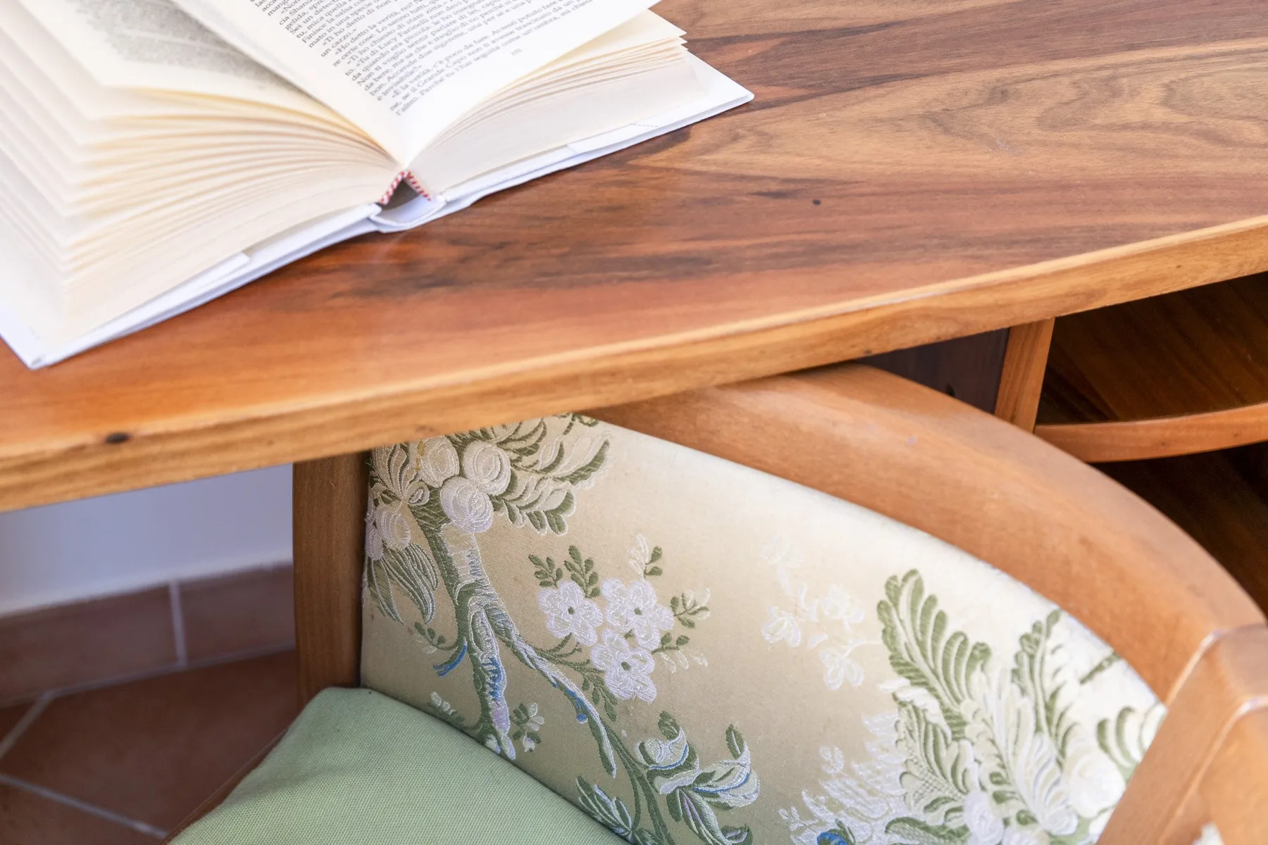 A wooden table with an open book sits above a floral upholstered chair.