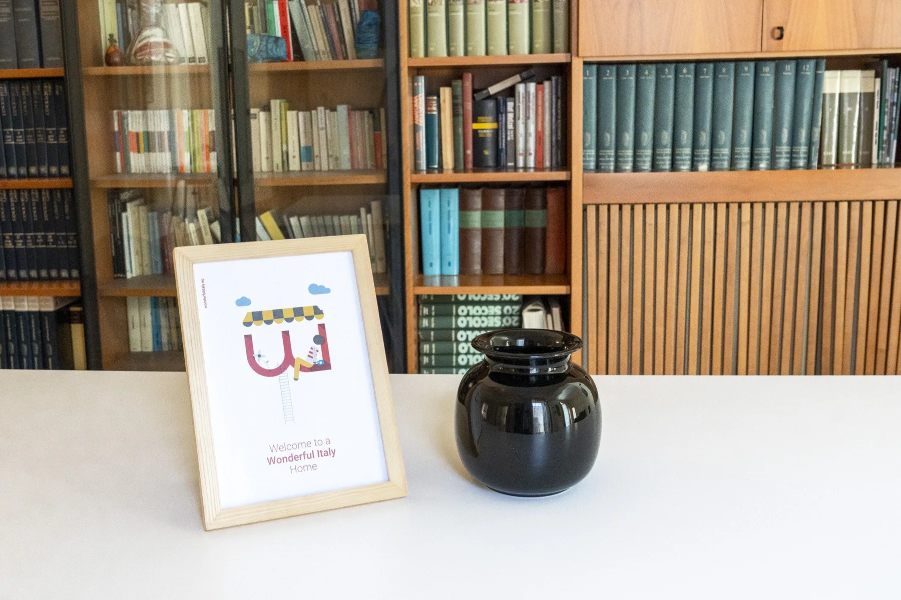 A framed print and a dark vase sit on a white table in front of bookshelves.