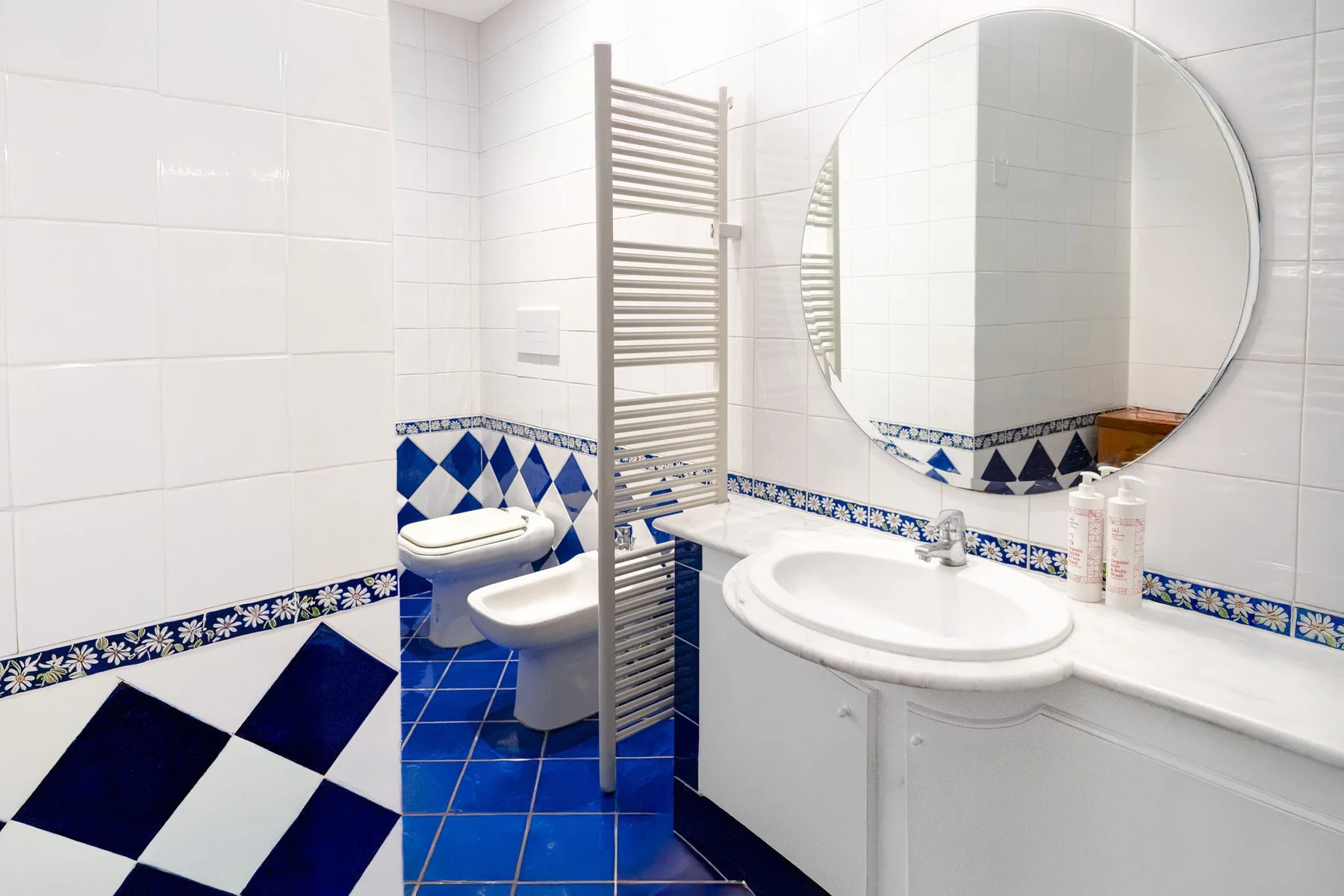 A tiled bathroom with a sink, round mirror, and towel warmer.