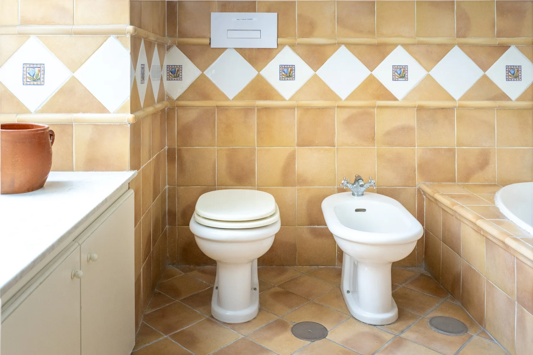 A tiled bathroom with a toilet and bidet beside a white countertop.
