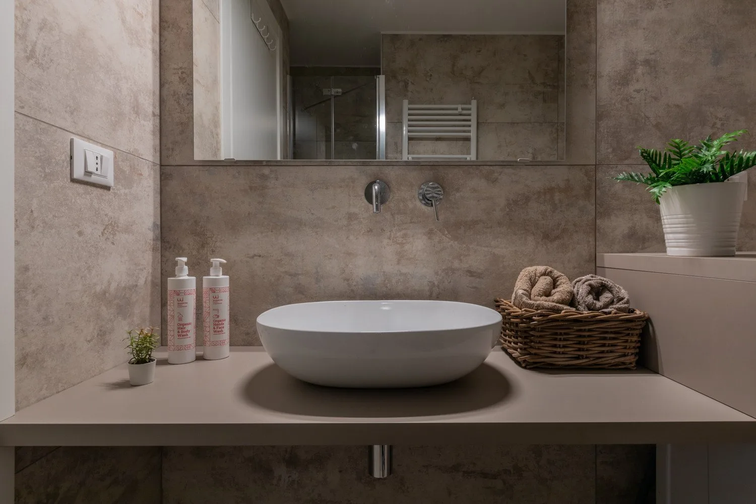 Un tocador de baño con lavabo sobre encimera, artículos de aseo y una cesta junto al espejo.