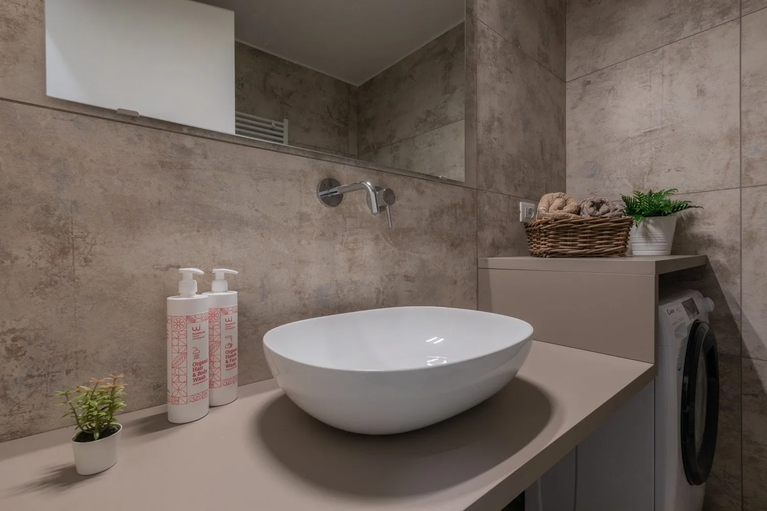 Un mueble de baño con lavabo blanco, grifo de pared y lavadora.