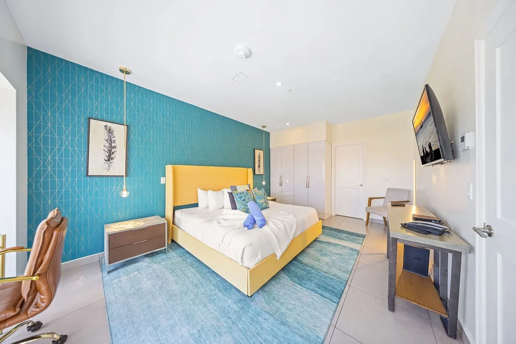 Your sun-soaked bedroom invites you to unwind with its vibrant teal accent wall, plush bed, and stylish decor. Bright, airy, and filled with coastal charm—your perfect retreat awaits.
