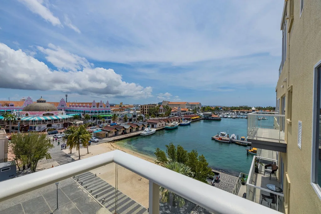 Colorful waterfront district with marina, shops, and tropical architecture visible from property balcony. Palm-lined streets and turquoise harbor waters frame the vibrant coastal scene.