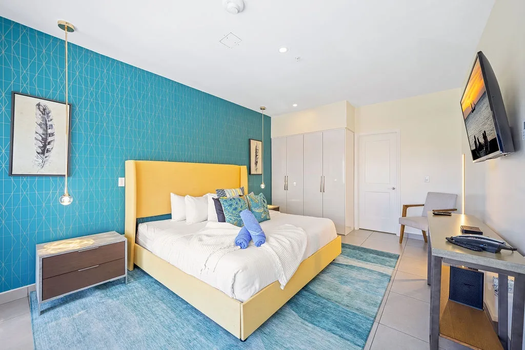 Unwind in your vibrant bedroom featuring a plush yellow bed, bold turquoise accent wall, and modern amenities like a wall-mounted TV and mini-fridge for your convenience.