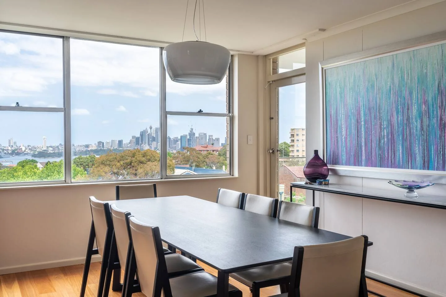 Una mesa de comedor está junto a grandes ventanales con vistas al agua y al perfil urbano.