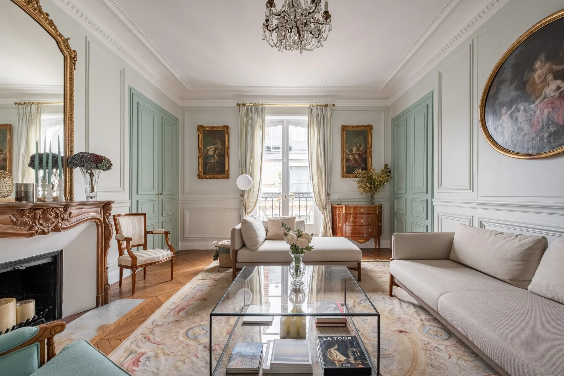 An elegant living room with pale seating, a glass coffee table, and tall windows.