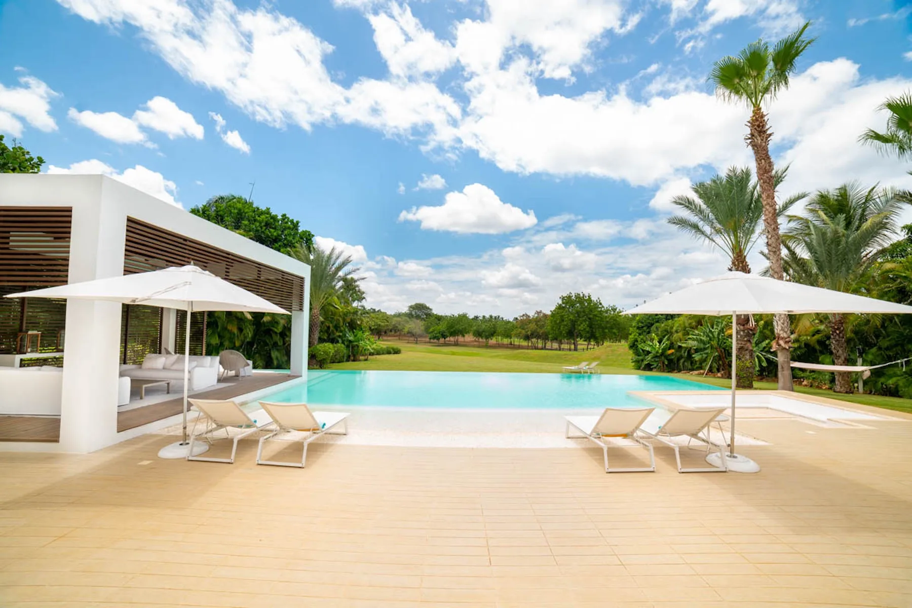Un patio junto a la piscina con asientos reclinables y sombrillas da a un paisaje verde.