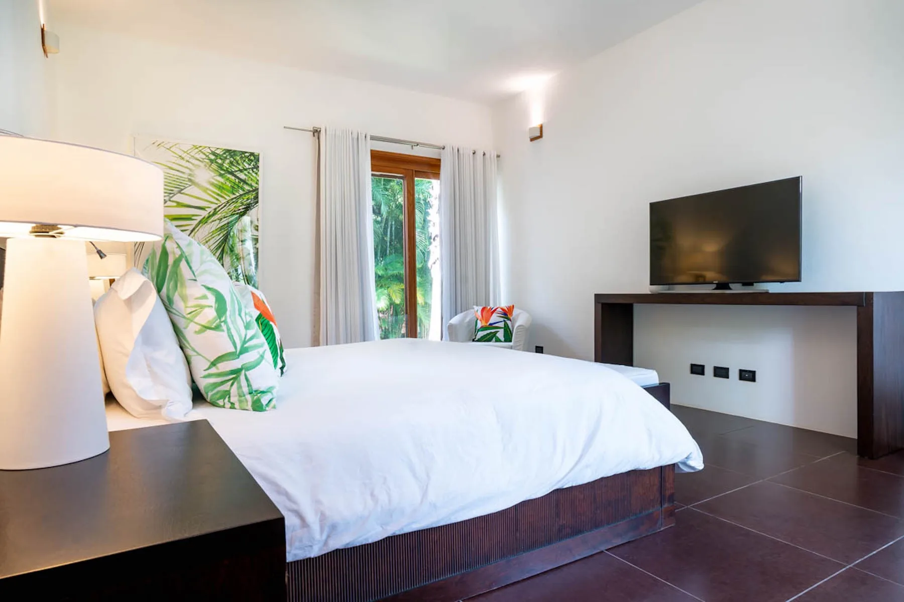 Un dormitorio luminoso con una cama blanca, una lámpara de noche y un televisor.