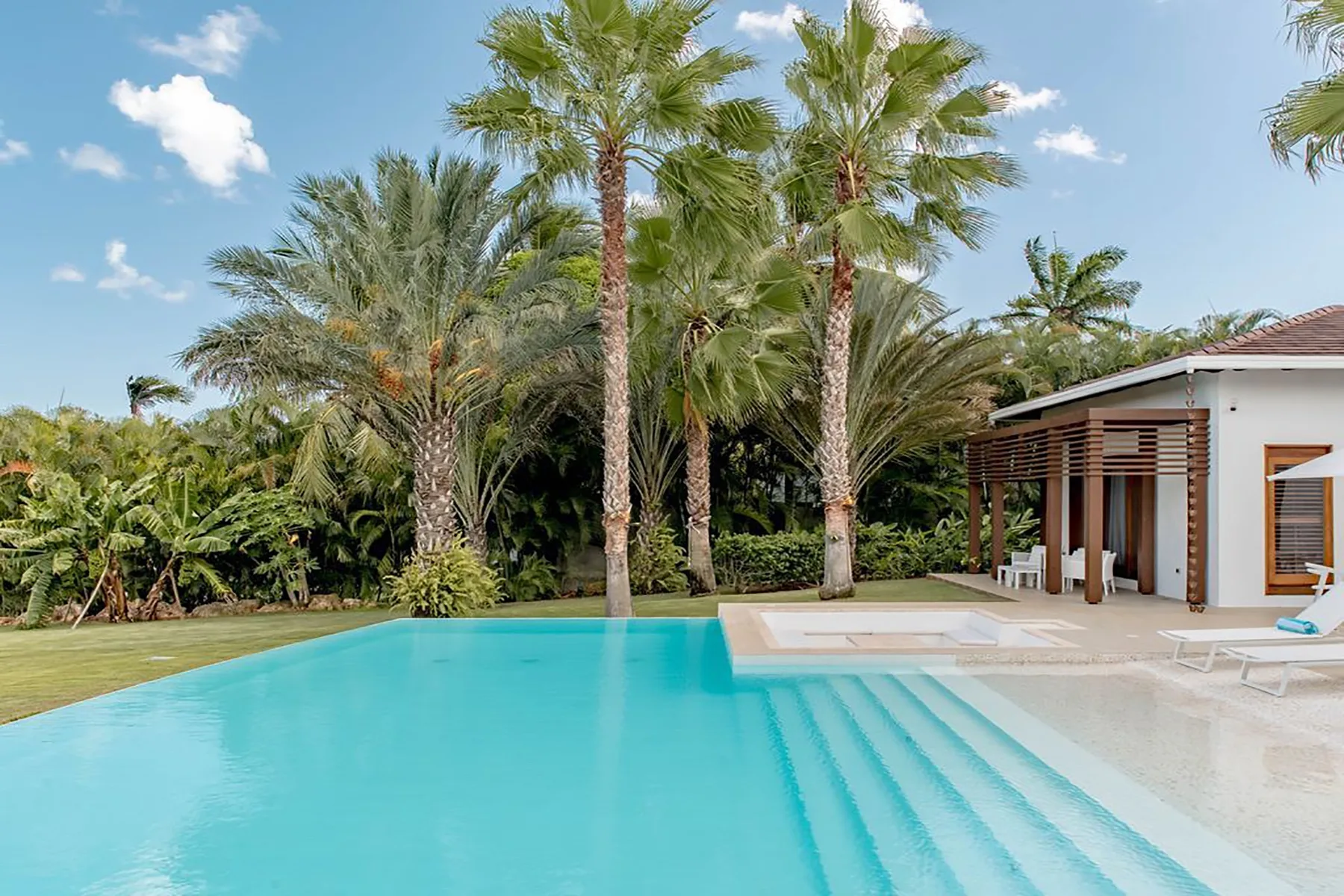 Una piscina junto a un patio, con palmeras y una casa al fondo.