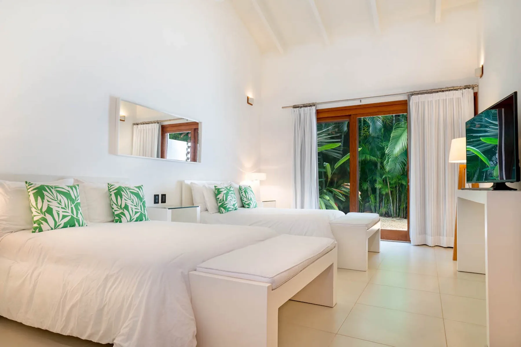 Un dormitorio luminoso con ropa de cama blanca, cojines verdes y puertas de vidrio frente a la vegetación.