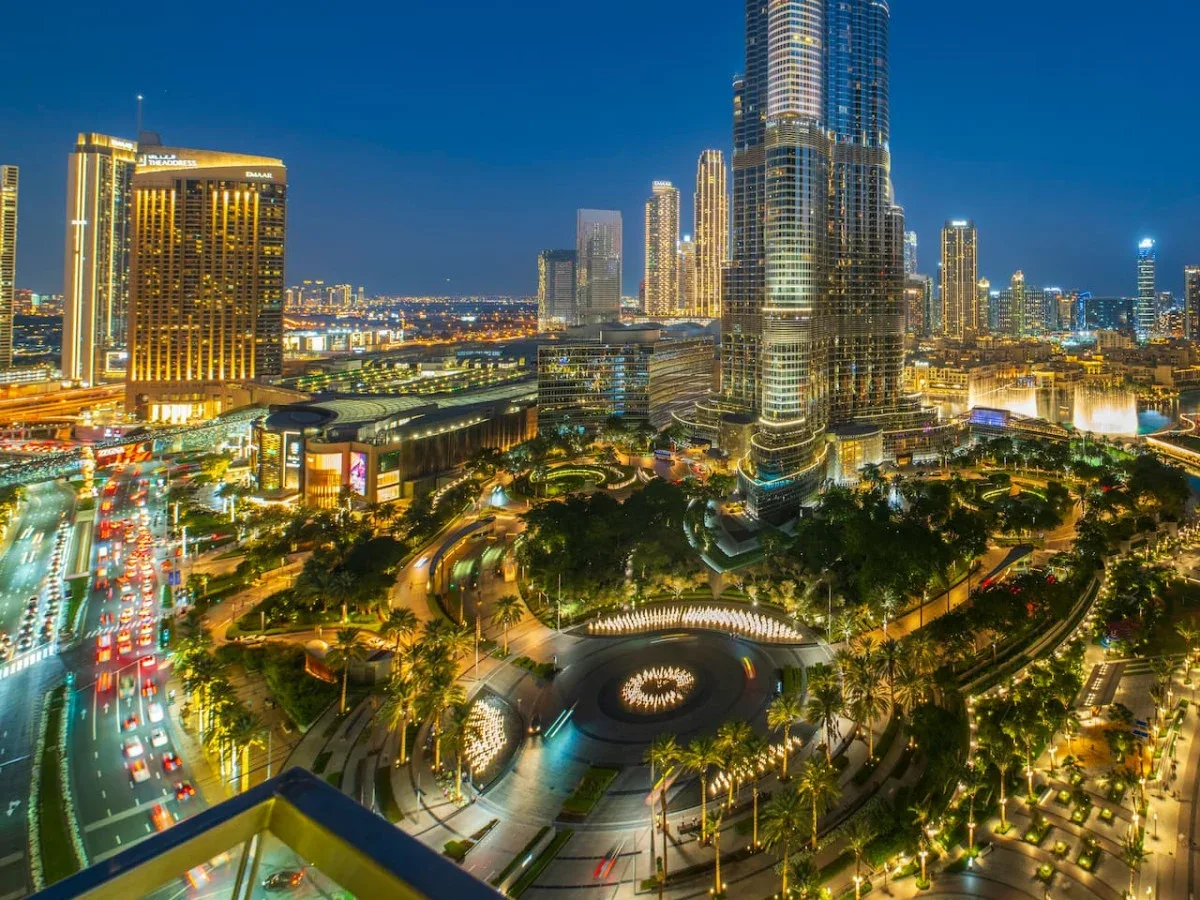 El horizonte nocturno de la ciudad domina una plaza iluminada y zonas ajardinadas.