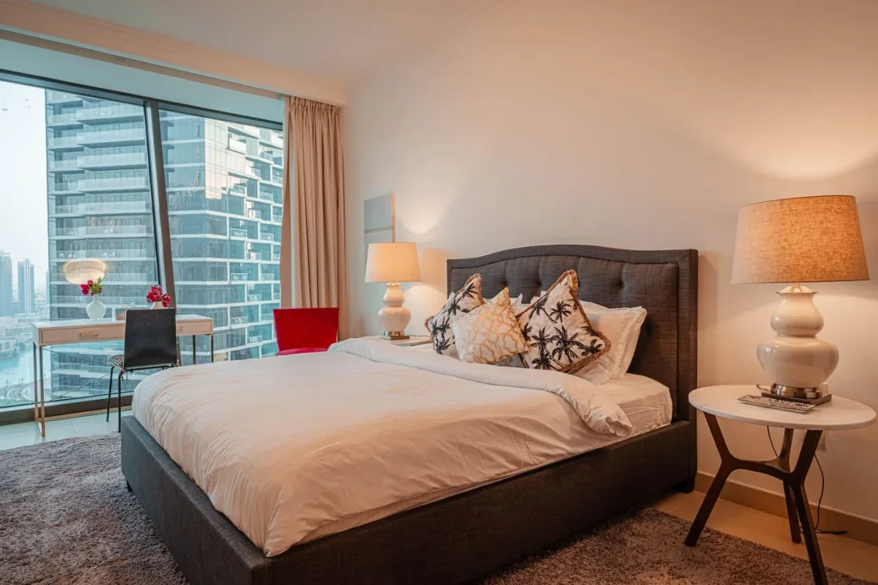 Un dormitorio con una cama grande junto a ventanales con vistas a edificios de la ciudad.