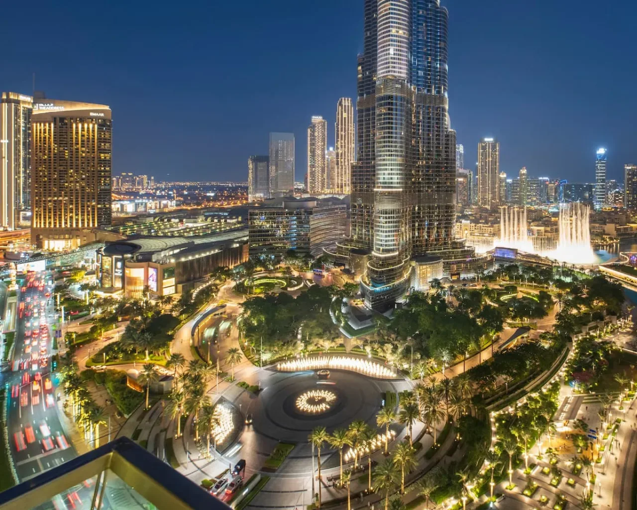 Un horizonte urbano iluminado de noche se alza sobre jardines y una plaza circular.