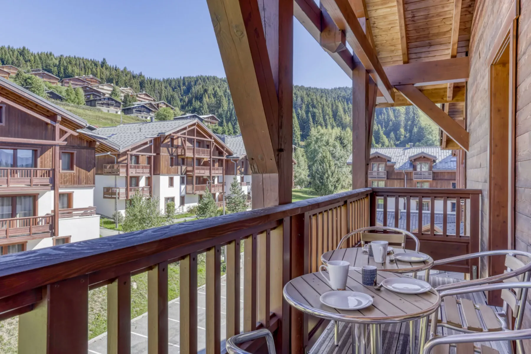 A covered wooden balcony with outdoor seating overlooks nearby buildings and a forested hillside.
