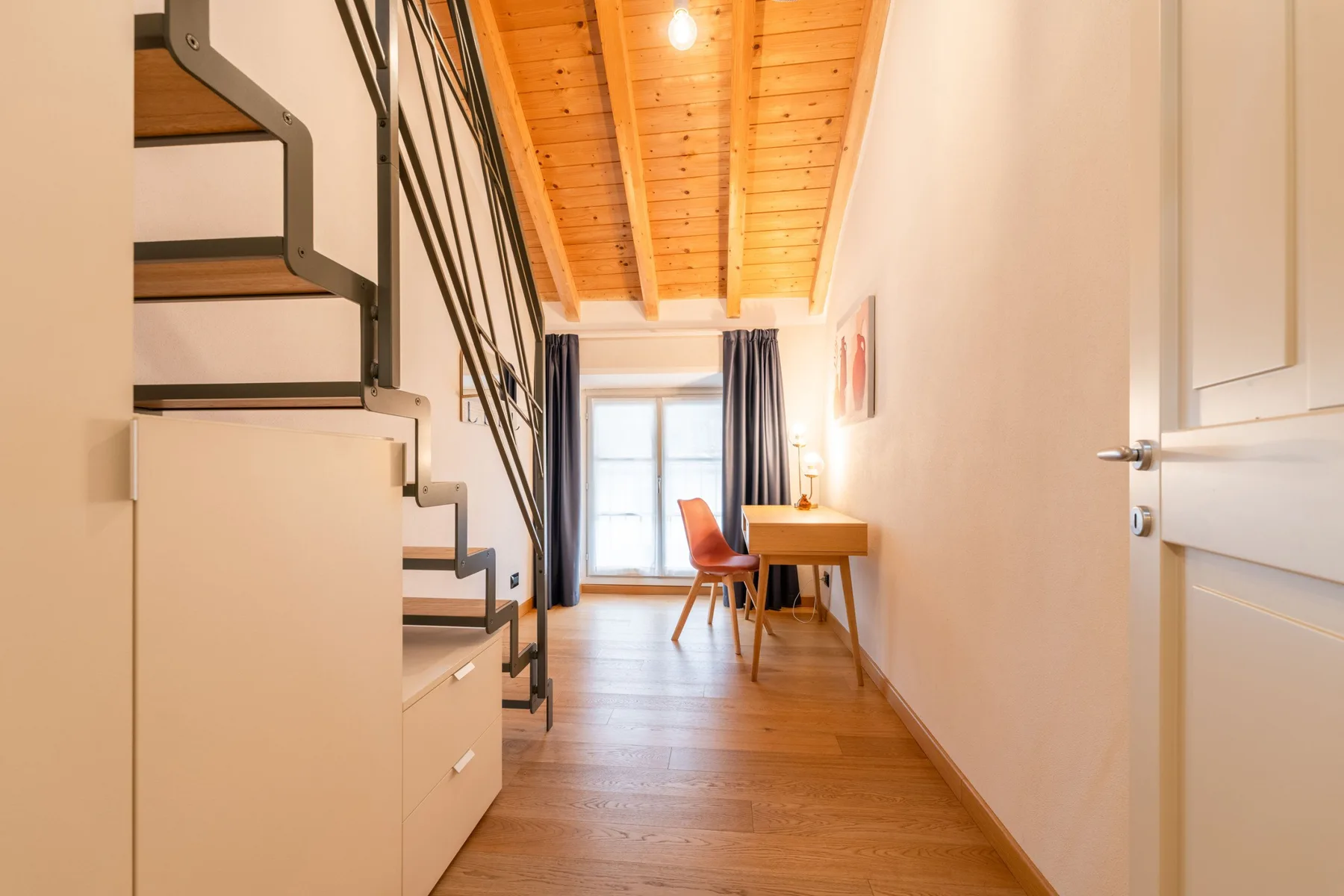 Un pasillo luminoso con una escalera, techo de madera y escritorio junto a la ventana.