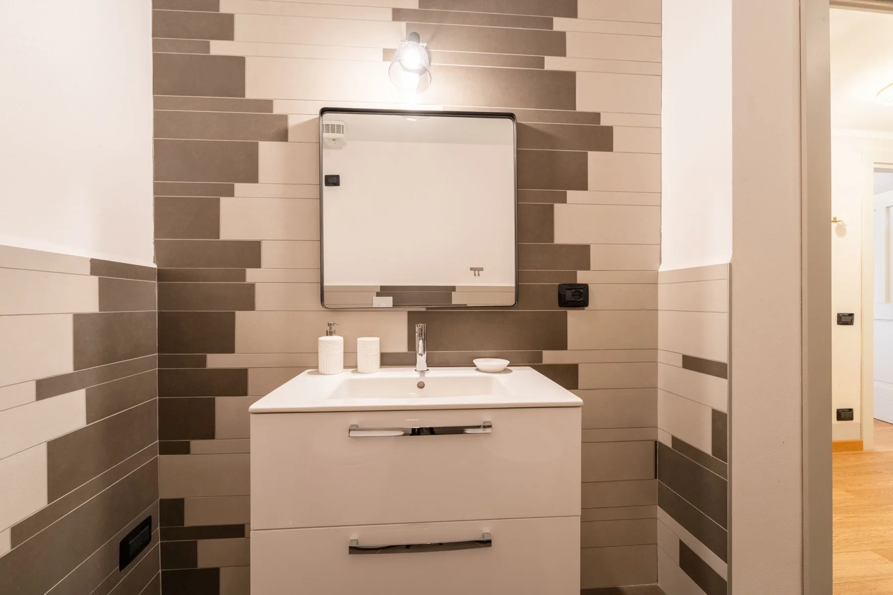 Un lavabo de baño con espejo frente a una pared de azulejos estampados.