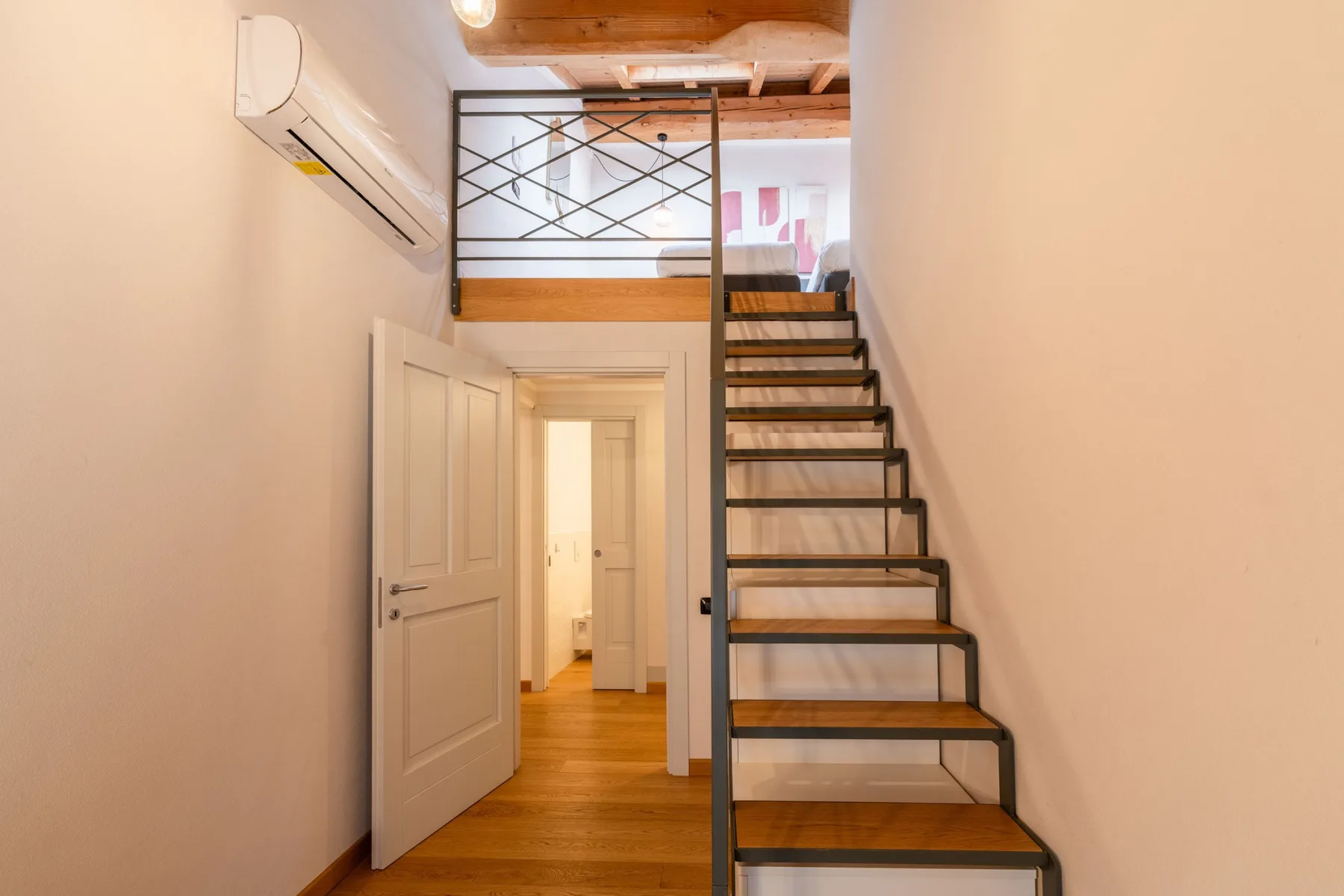 Una escalera de madera sube junto a una puerta abierta en un interior luminoso.
