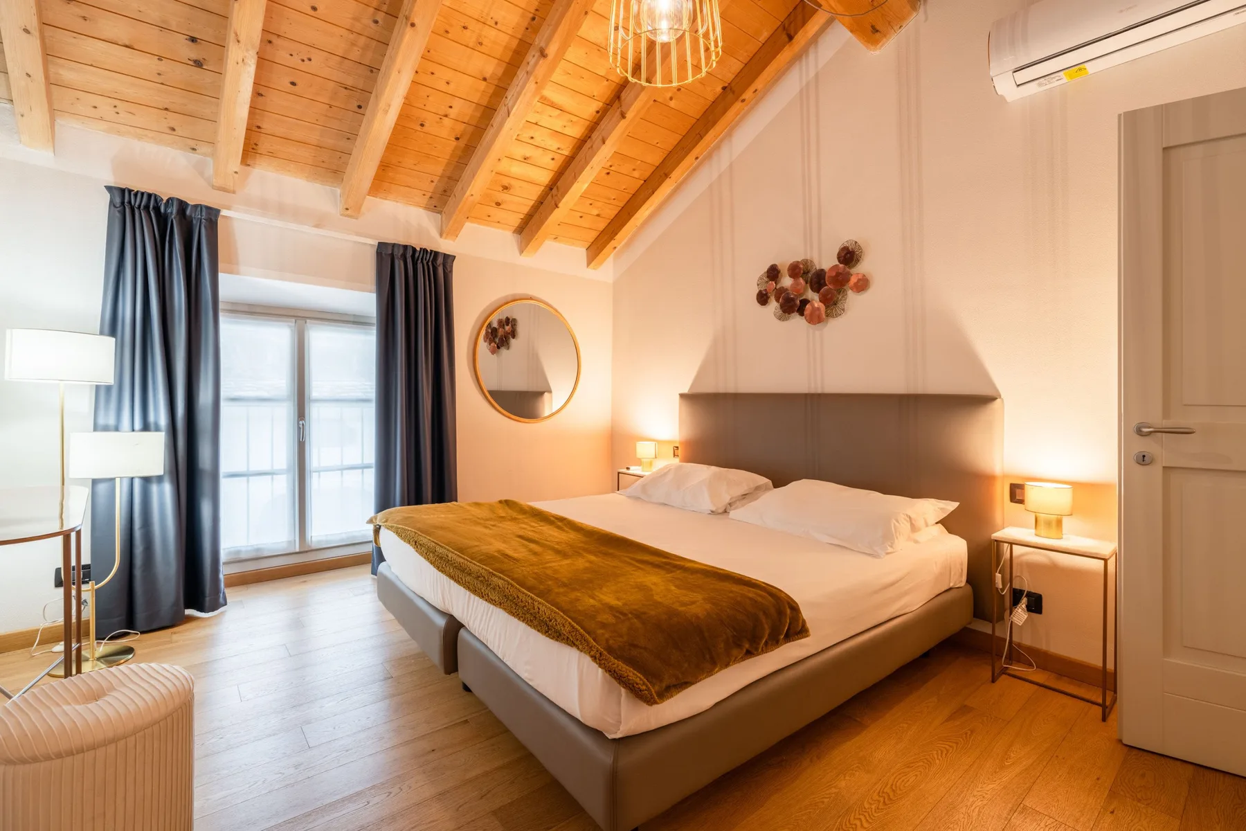 Un dormitorio con una cama bajo un techo de madera inclinado y ventanas con cortinas oscuras.