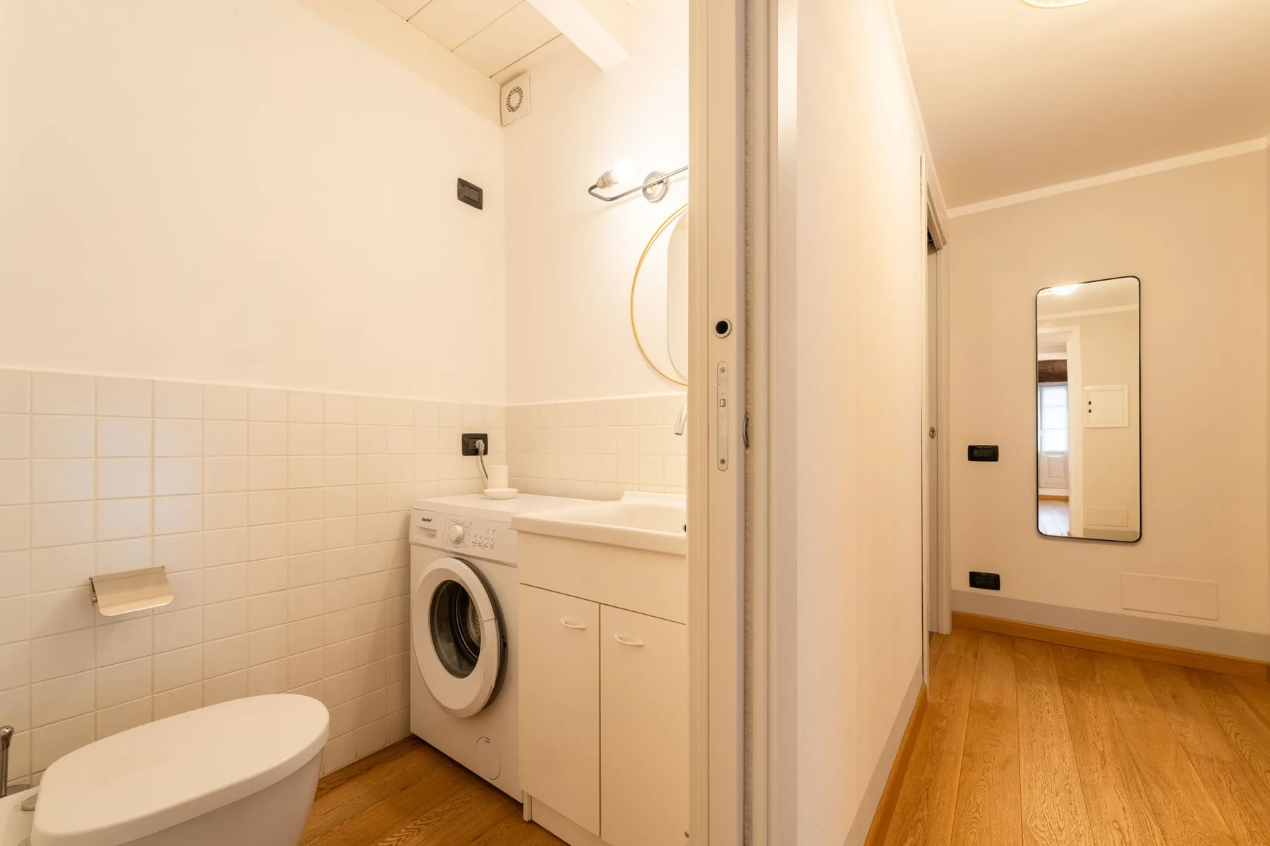 Un baño con inodoro, lavadora y lavabo junto a un pasillo con suelo de madera.