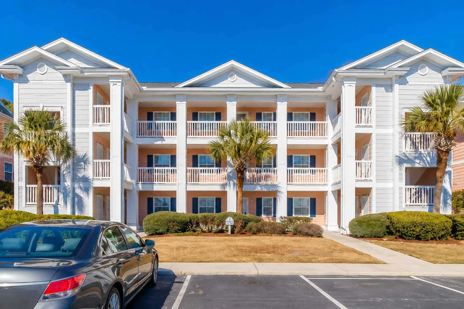 A white multi-story building with balconies stands beside a parking area.