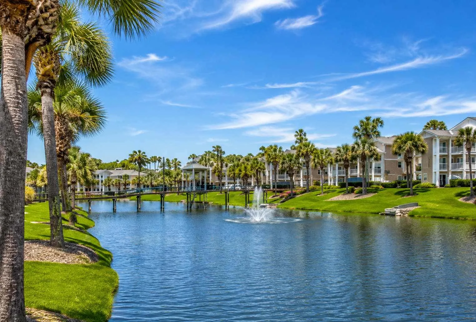 A landscaped pond with a fountain and palm trees beside residential buildings.
