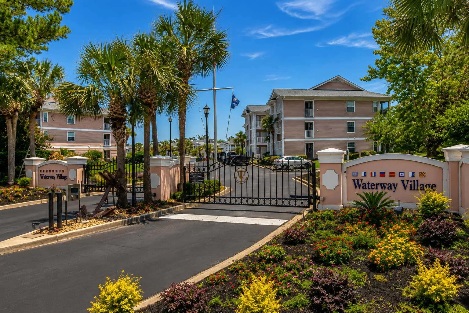 A gated residential community entrance with landscaping and apartment buildings beyond.