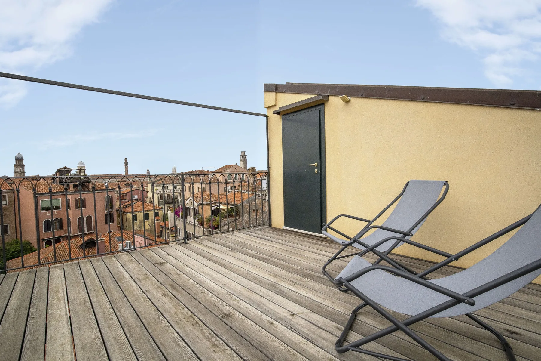 Una terraza en la azotea con suelo de madera, asientos reclinables y una barandilla metálica frente a edificios cercanos.