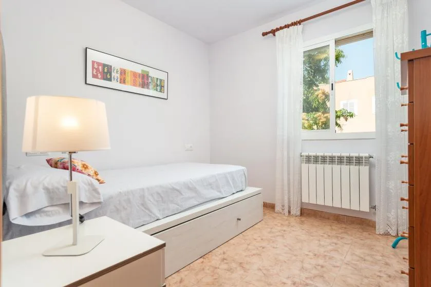 Un dormitorio luminoso con una cama individual, una lámpara de mesilla y una ventana con cortinas translúcidas.