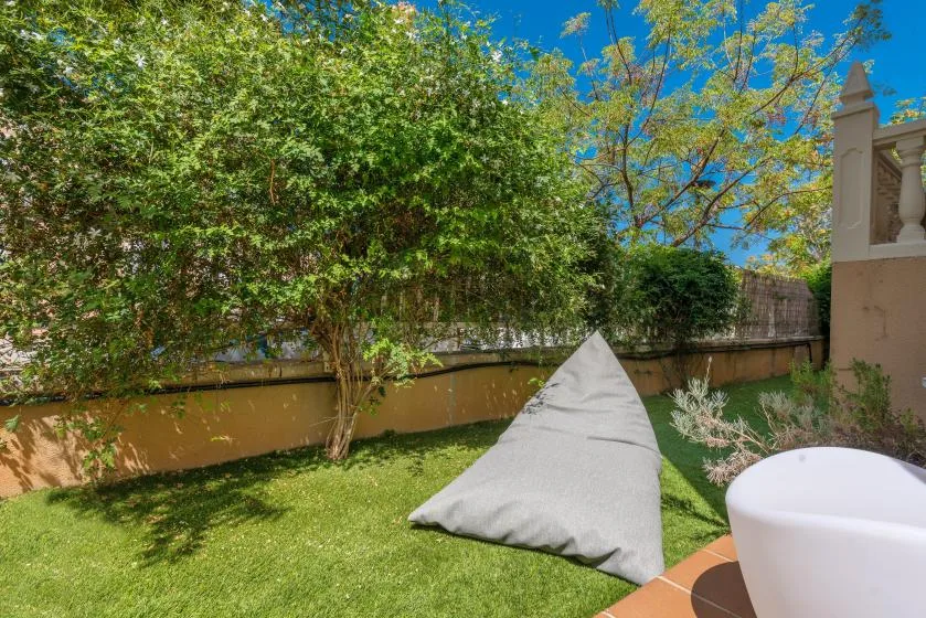 Un jardín con césped, árboles frondosos y un cojín gris de exterior.