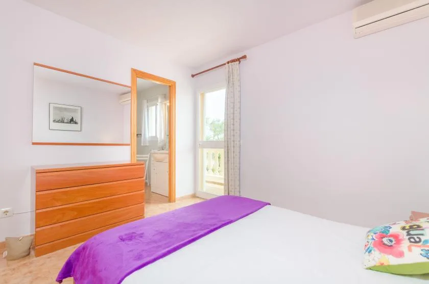 Un dormitorio luminoso con una cama, una colcha morada y una cómoda de madera.