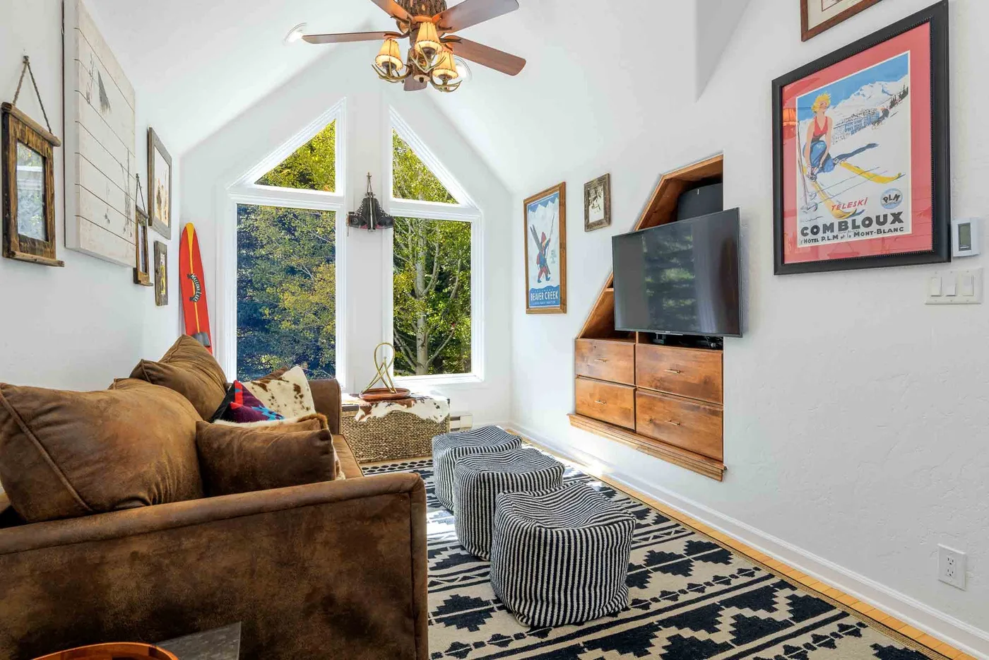 Una sala luminosa con un sofá marrón, televisor y ventanas grandes.